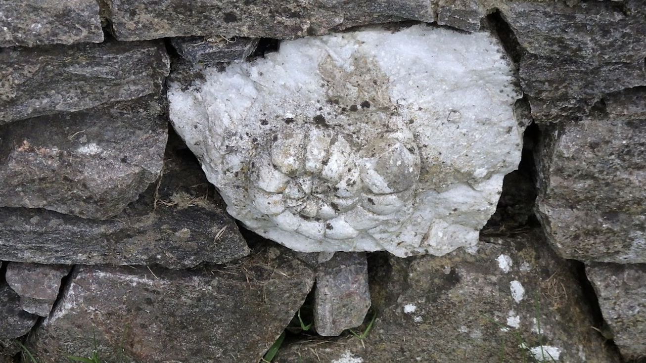 The image displays a large, circular ammonite fossil embedded in a stone wall. The fossil stands out due to its lighter color and distinctive ribbed spiral pattern, contrasting with the darker, rough-textured stones surrounding it